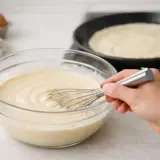 Préparation de la pâte à crêpe dans un bol en verre, avec une crêpe en cuisson dans une poêle au fond.