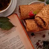 Crêpes roulées sur une assiette, accompagnées d'une tasse de thé et d'un livre de recette. Une framboise décore les crêpes. Parfait pour une recette crêpe.