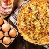 Une délicieuse quiche lorraine dorée, garnie de lardons et de ciboulette, accompagnée d'œufs frais et de lard fumé sur une planche en bois.