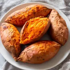 Cinq patates douces rôties à la perfection dans un air fryer, servies sur une assiette blanche. L'une est coupée en deux, révélant sa chair orange tendre.