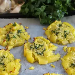 Cinq écrasés de pomme de terre dorés, garnis d'herbes et d'ail, sur du papier sulfurisé.