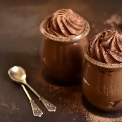 Deux verrines de recette mousse au chocolat au lait, saupoudrées de cacao, avec deux cuillères anciennes.