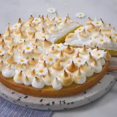 Une magnifique tarte aux citrons meringuée, décorée de petites fleurs blanches et jaunes, avec une part déjà coupée.