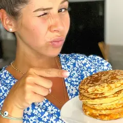 Une femme fait un clin d'œil et pointe du doigt une pile de pancakes banane sans oeuf sur une assiette.