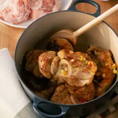 Jarret de veau mijoté dans une cocotte en fonte, garni d'herbes. Une cuillère en bois repose sur la viande.