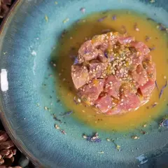 Un délicieux tartare de thon, parsemé de graines de sésame et de fleurs bleues, baignant dans une sauce dorée sur une assiette texturée.
