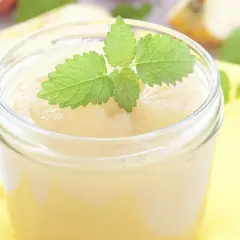 Un pot de compote pomme banane, garni d'une feuille de menthe fraîche. Des morceaux de pommes flous en arrière-plan.