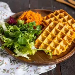 Gaufre patate douce dorée, servie avec une salade verte fraîche et une julienne de carottes râpées sur une assiette en bois.
