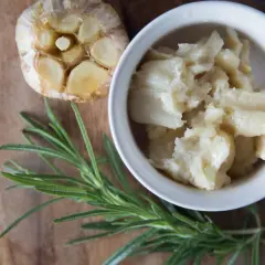 Ail confit au four, doux et crémeux, servi dans un bol blanc, à côté d'une tête d'ail rôtie et d'une branche de romarin frais.
