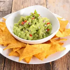 Un bol de guacamole traditionnel, garni de coriandre, est servi avec des chips tortilla sur une assiette blanche.