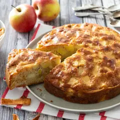 Un délicieux gâteau aux pommes, doré et moelleux, avec des morceaux de fruits visibles. Des pommes fraîches et des chips de pommes complètent cette scène gourmande.