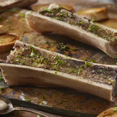 Deux os à moelle rôtis, parsemés de persil et de poivre, accompagnés de toasts grillés. Une cuillère attend patiemment.