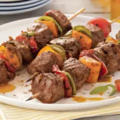 Brochettes de bœuf marinées, garnies de poivrons et tomates, sur une assiette blanche.