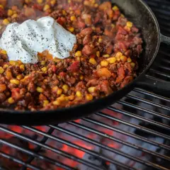 Chili con carne fumant dans une poêle en fonte sur un barbecue, garni d'une cuillère de crème fraîche poivrée.