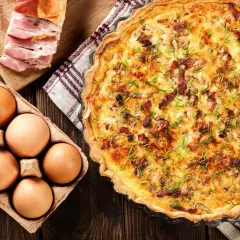 Une délicieuse quiche lorraine dorée, garnie de lardons et de ciboulette, accompagnée d'œufs frais et de lard fumé sur une planche en bois.