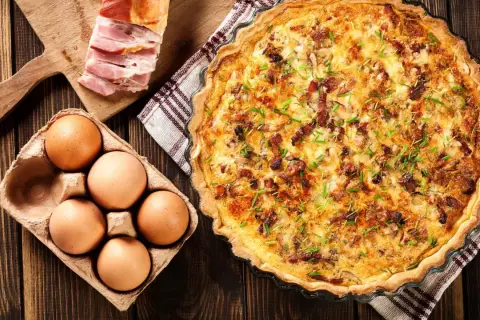 Une délicieuse quiche lorraine dorée, garnie de lardons et de ciboulette, accompagnée d'œufs frais et de lard fumé sur une planche en bois.