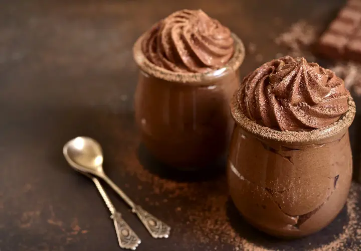 Deux verrines de recette mousse au chocolat au lait, saupoudrées de cacao, avec deux cuillères anciennes.