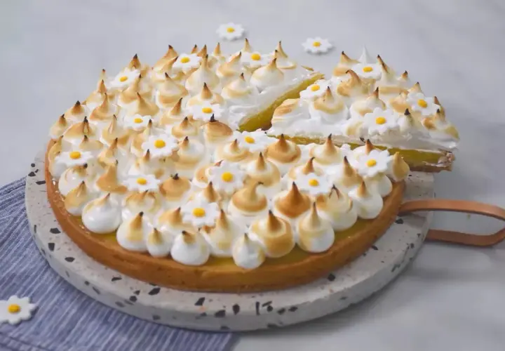 Une magnifique tarte aux citrons meringuée, décorée de petites fleurs blanches et jaunes, avec une part déjà coupée.