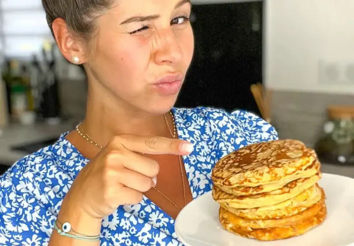 Une femme fait un clin d'œil et pointe du doigt une pile de pancakes banane sans oeuf sur une assiette.