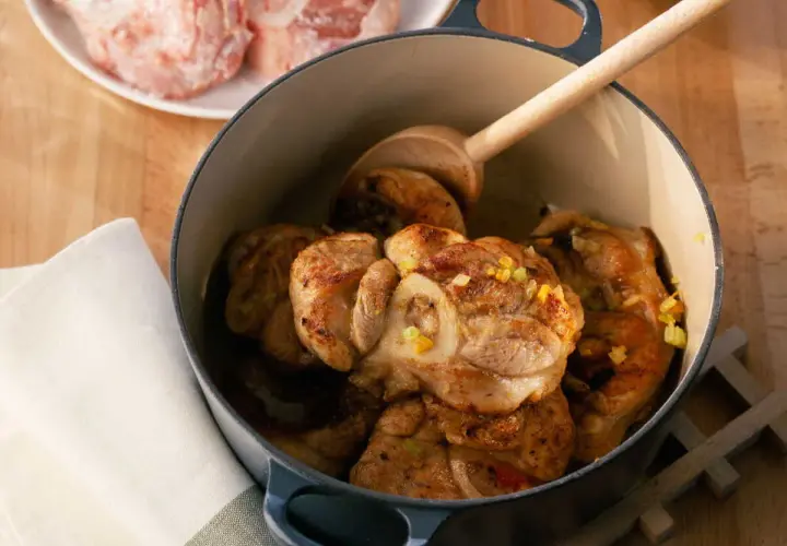 Jarret de veau mijoté dans une cocotte en fonte, garni d'herbes. Une cuillère en bois repose sur la viande.