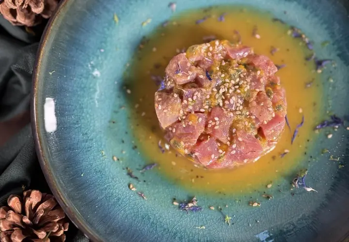 Un délicieux tartare de thon, parsemé de graines de sésame et de fleurs bleues, baignant dans une sauce dorée sur une assiette texturée.
