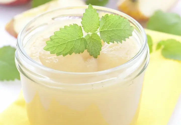 Un pot de compote pomme banane, garni d'une feuille de menthe fraîche. Des morceaux de pommes flous en arrière-plan.
