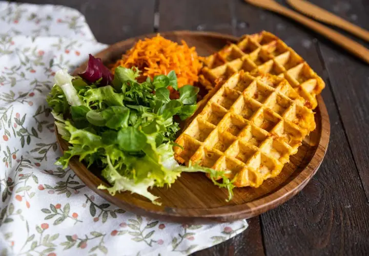 Gaufre patate douce dorée, servie avec une salade verte fraîche et une julienne de carottes râpées sur une assiette en bois.