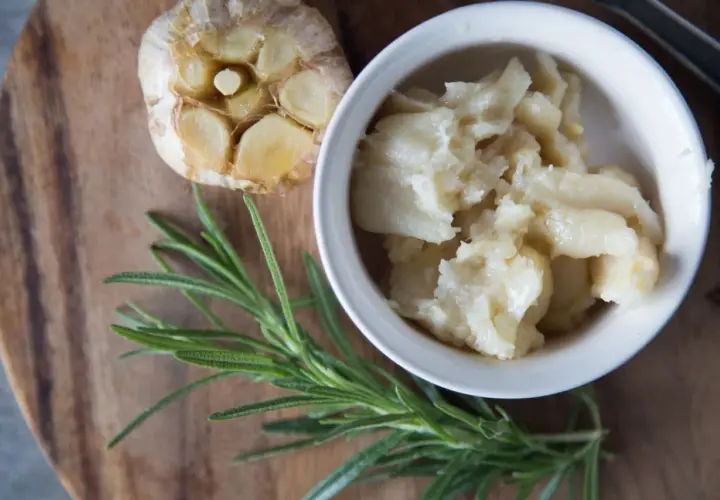 Ail confit au four, doux et crémeux, servi dans un bol blanc, à côté d'une tête d'ail rôtie et d'une branche de romarin frais.