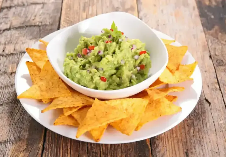 Un bol de guacamole traditionnel, garni de coriandre, est servi avec des chips tortilla sur une assiette blanche.