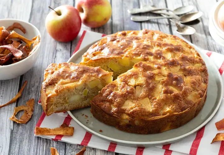 Un délicieux gâteau aux pommes, doré et moelleux, avec des morceaux de fruits visibles. Des pommes fraîches et des chips de pommes complètent cette scène gourmande.