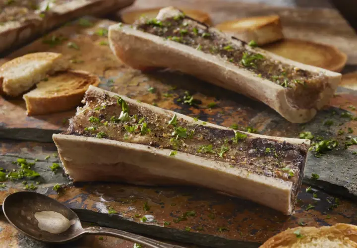Deux os à moelle rôtis, parsemés de persil et de poivre, accompagnés de toasts grillés. Une cuillère attend patiemment.