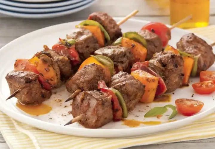 Brochettes de bœuf marinées, garnies de poivrons et tomates, sur une assiette blanche.