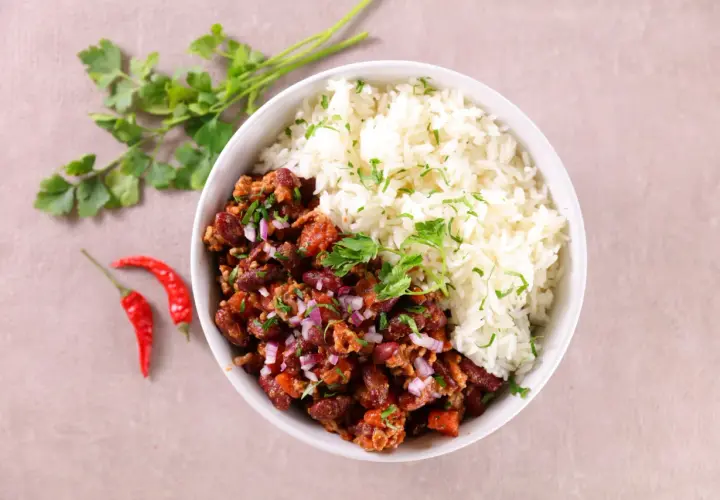 Un bol de chili con carne et riz, garni de persil frais et d'oignons rouges. Deux piments rouges sont posés à côté.