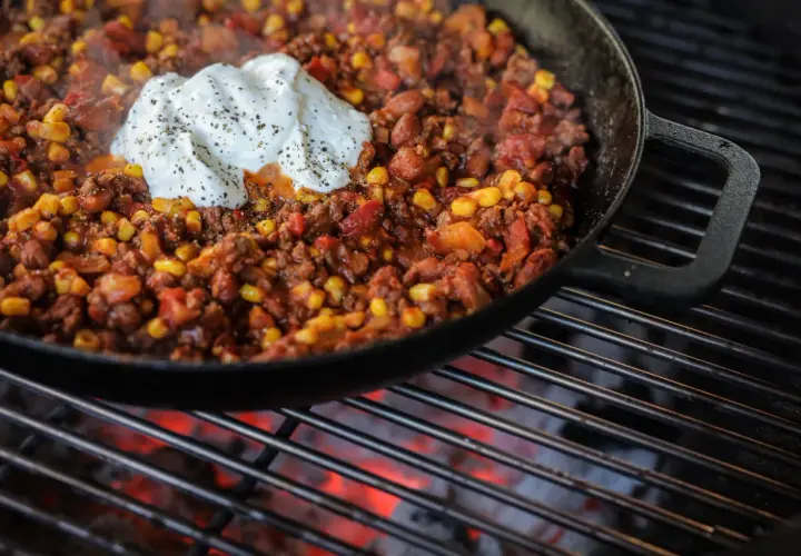 Chili con carne fumant dans une poêle en fonte sur un barbecue, garni d'une cuillère de crème fraîche poivrée.