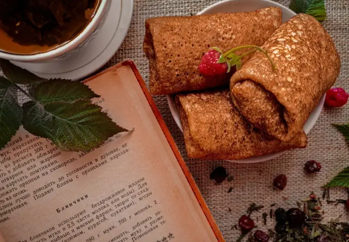 Crêpes roulées sur une assiette, accompagnées d'une tasse de thé et d'un livre de recette. Une framboise décore les crêpes. Parfait pour une recette crêpe.