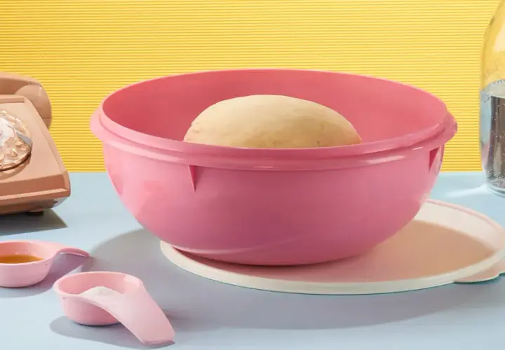 Pâte à pain dans un bol Tupperware rose, prêt pour une recette minute. À côté, un téléphone vintage, des cuillères doseuses et une bouteille d'eau.