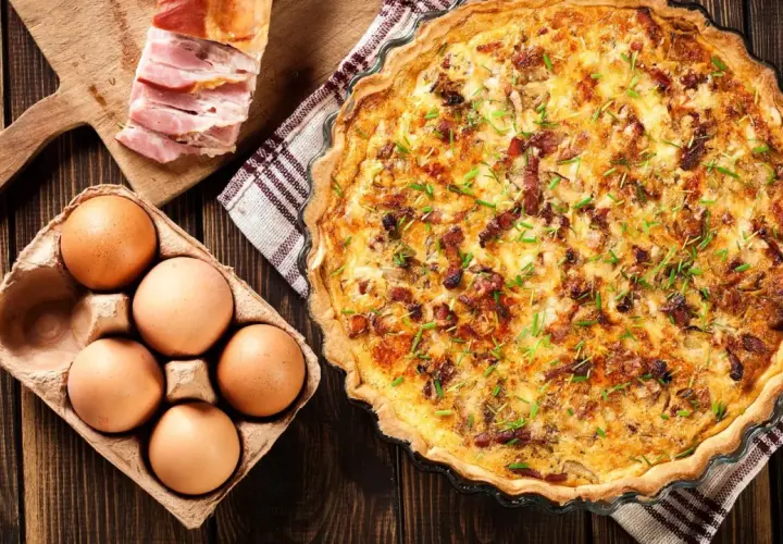 Une délicieuse quiche lorraine dorée, garnie de lardons et de ciboulette, accompagnée d'œufs frais et de lard fumé sur une planche en bois.