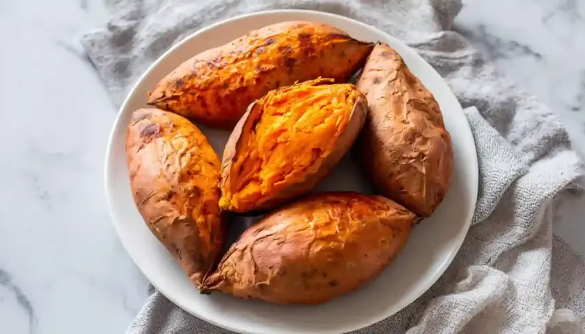 Cinq patates douces rôties à la perfection dans un air fryer, servies sur une assiette blanche. L'une est coupée en deux, révélant sa chair orange tendre.