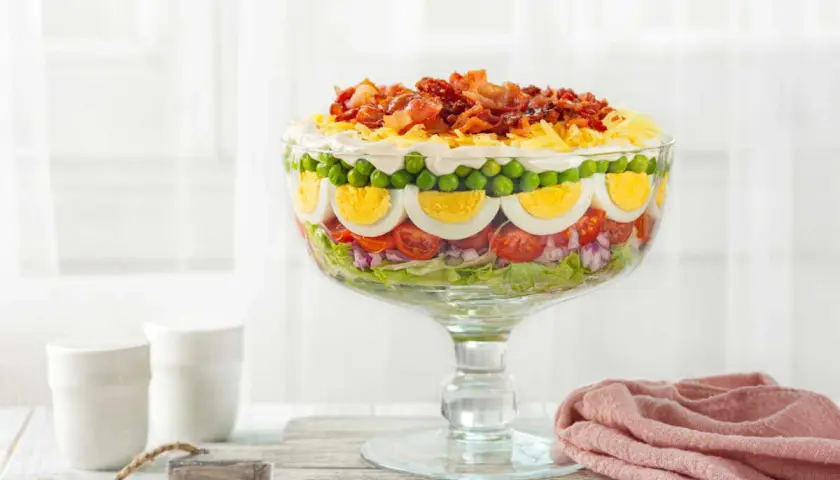 Une magnifique salade 7 étages, présentée dans un grand bol en verre, avec des couches de légumes frais, d'œufs durs, de petits pois et de bacon croustillant sur le dessus.