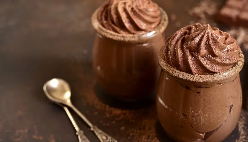 Deux verrines de recette mousse au chocolat au lait, saupoudrées de cacao, avec deux cuillères anciennes.