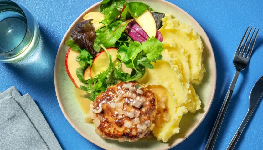 Un steak haché sauce oignons, servi avec une généreuse portion de purée de pomme de terre et une salade croquante aux pommes.