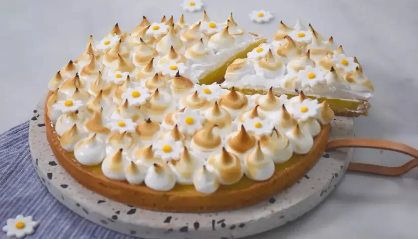 Une magnifique tarte aux citrons meringuée, décorée de petites fleurs blanches et jaunes, avec une part déjà coupée.