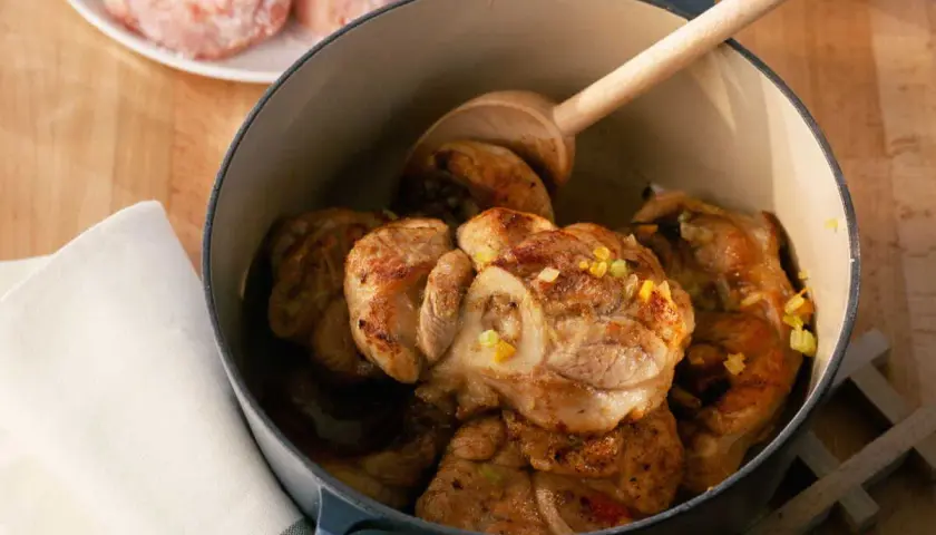 Jarret de veau mijoté dans une cocotte en fonte, garni d'herbes. Une cuillère en bois repose sur la viande.