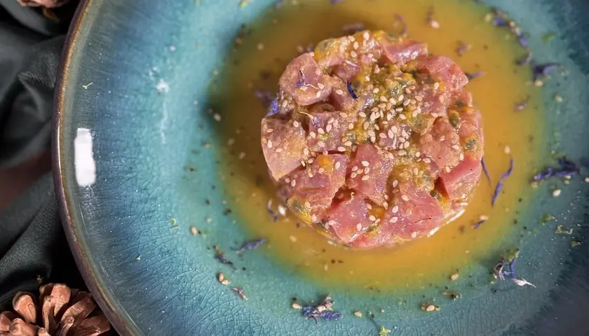 Un délicieux tartare de thon, parsemé de graines de sésame et de fleurs bleues, baignant dans une sauce dorée sur une assiette texturée.