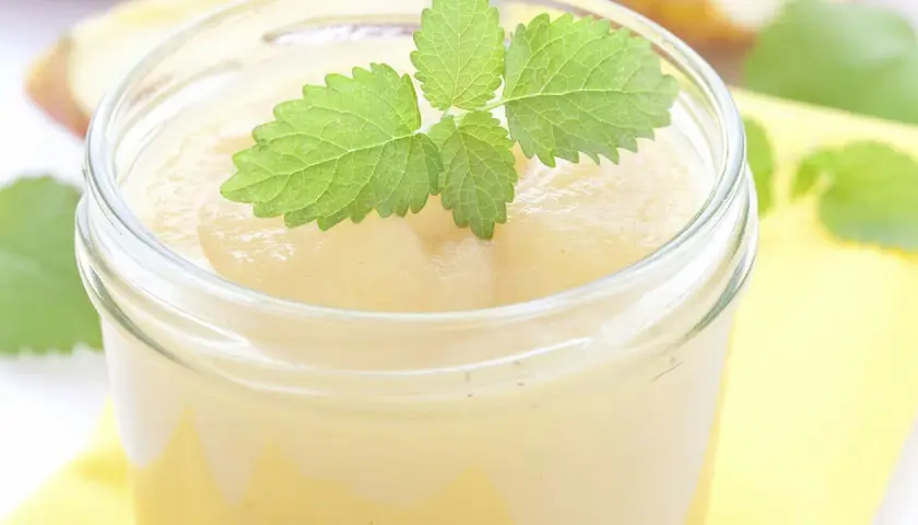 Un pot de compote pomme banane, garni d'une feuille de menthe fraîche. Des morceaux de pommes flous en arrière-plan.