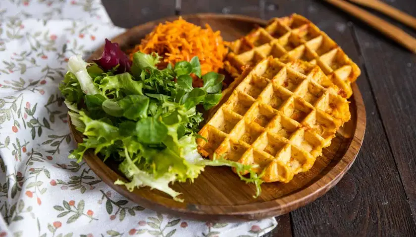 Gaufre patate douce dorée, servie avec une salade verte fraîche et une julienne de carottes râpées sur une assiette en bois.