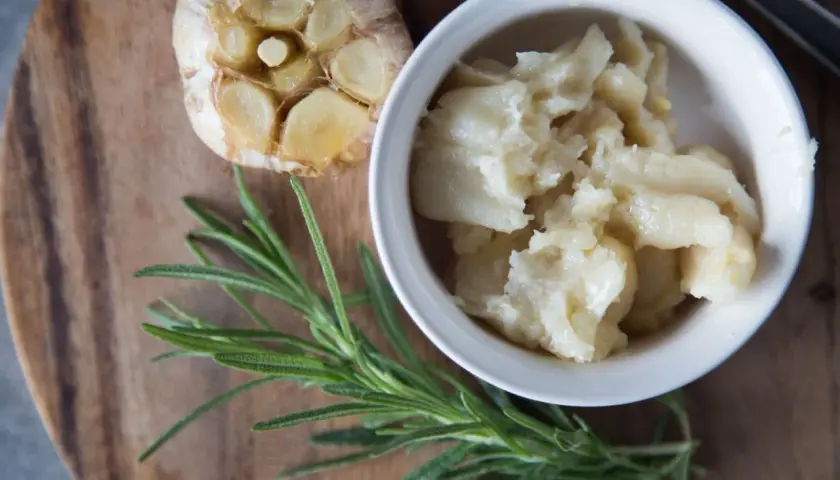 Ail confit au four, doux et crémeux, servi dans un bol blanc, à côté d'une tête d'ail rôtie et d'une branche de romarin frais.
