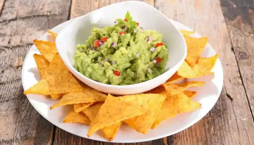 Un bol de guacamole traditionnel, garni de coriandre, est servi avec des chips tortilla sur une assiette blanche.