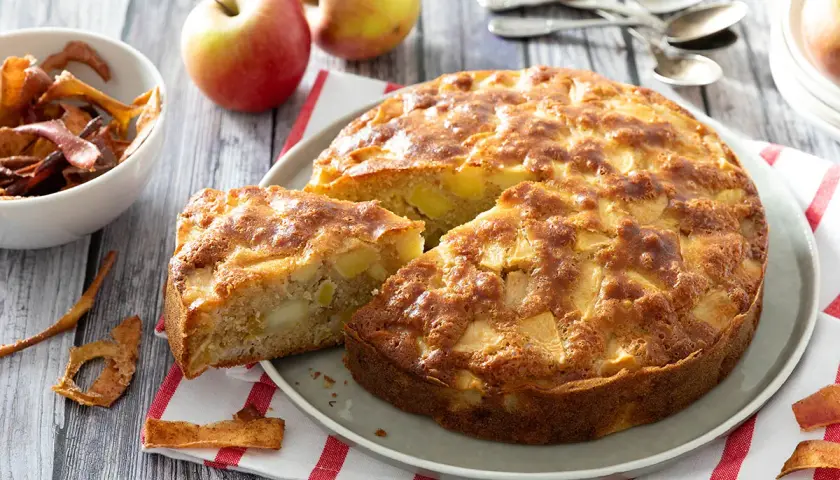 Un délicieux gâteau aux pommes, doré et moelleux, avec des morceaux de fruits visibles. Des pommes fraîches et des chips de pommes complètent cette scène gourmande.