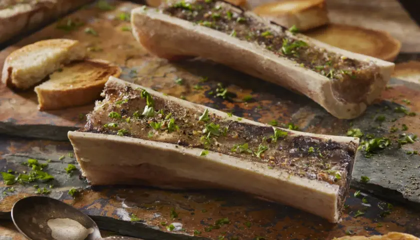 Deux os à moelle rôtis, parsemés de persil et de poivre, accompagnés de toasts grillés. Une cuillère attend patiemment.